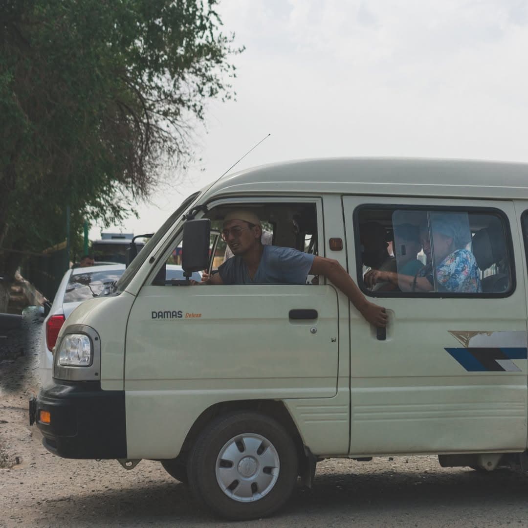 A marshrutka driver leans out of a white Damas minivan with his hand on the passenger door, on a dusty street in Nukus.