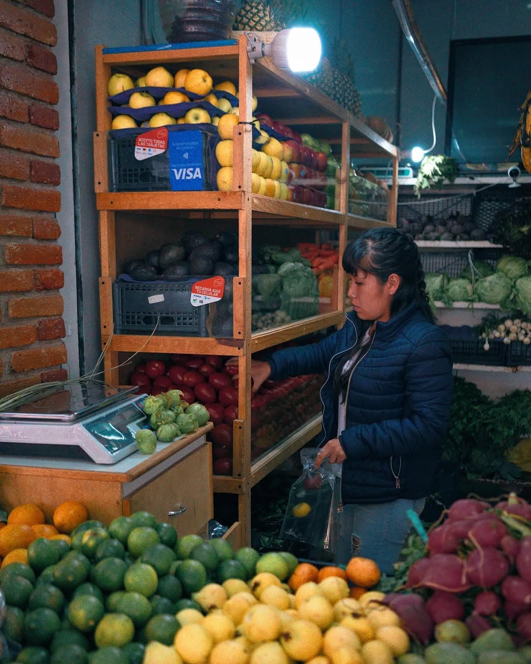 A woman selects tomatoes from wooden shelves stacked with fruit and vegetables at a small market stall.
