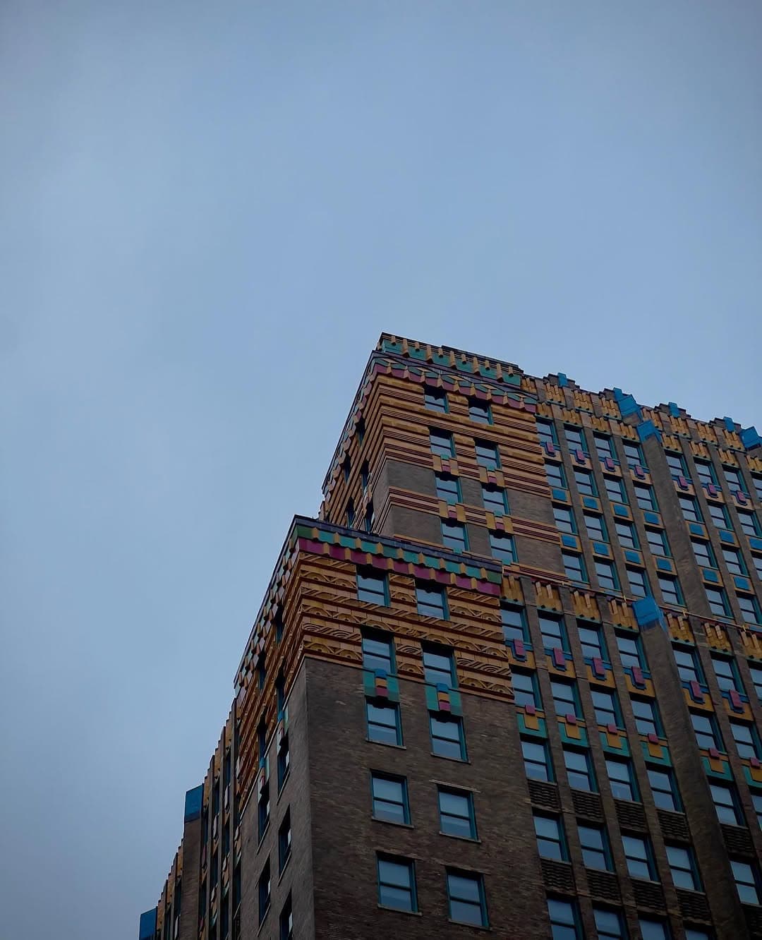 Looking up at the colorful Art Deco ornament on the upper floors of a brick building against an evening sky.