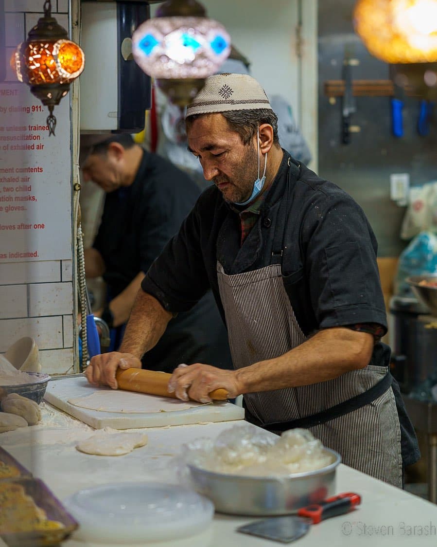 A cook in a skullcap and apron rolls out dough on a floured counter.