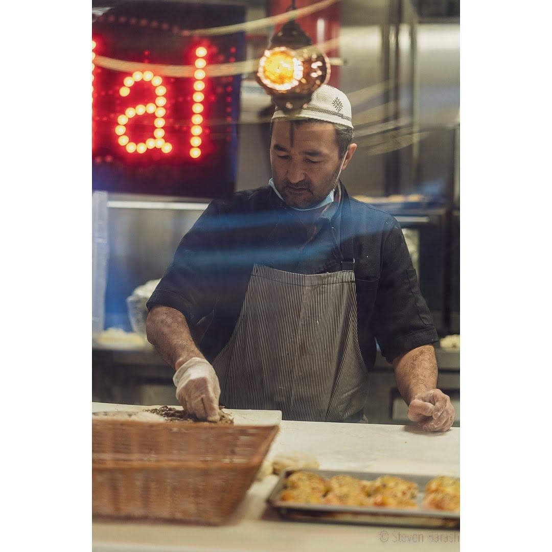 Seen through the restaurant window, a cook prepares food at a counter beside a tray of baked goods, under a red neon sign.