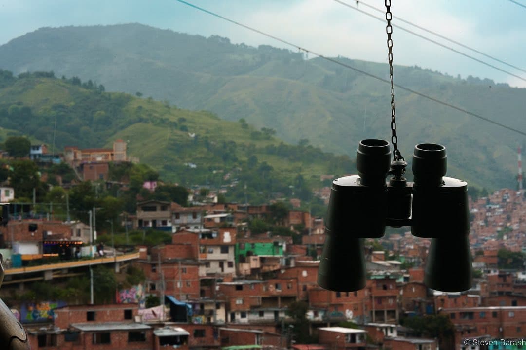 Binoculars hang in the foreground over the brick hillside homes of Medellín, with green mountains behind them.