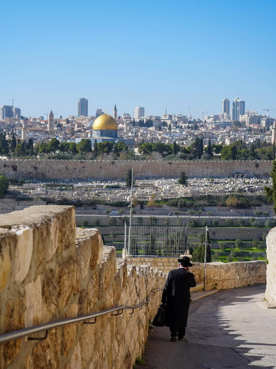 A man in a black hat and coat walks a path on the Mount of Olives, overlooking Jerusalem’s Old City walls and the Dome of the Rock.