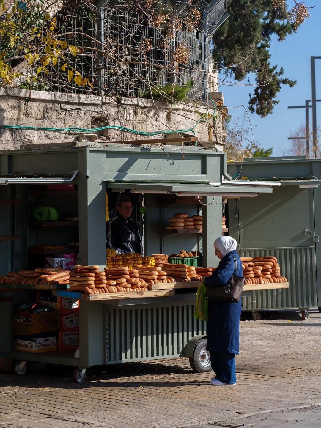 A woman in a white hijab buys bread from a street cart stacked with ka’ak rings outside Damascus Gate.
